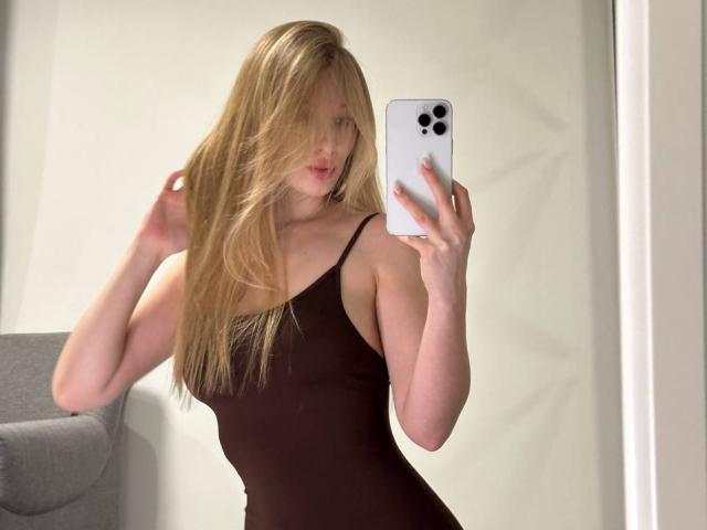 StellaLovely with long blonde hair, wearing a black spaghetti-strap dress, taking a mirror selfie with a white iPhone, showing her tits and a subtle ass curve.