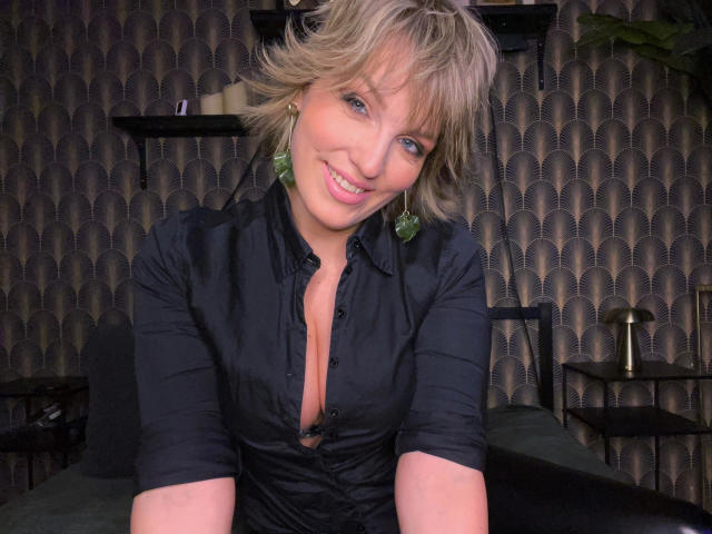 CharleneAngel-hot with short blonde hair, blue eyes, green earrings, smiling, wearing a black shirt with a deep cleavage, sitting in a dimly lit room.