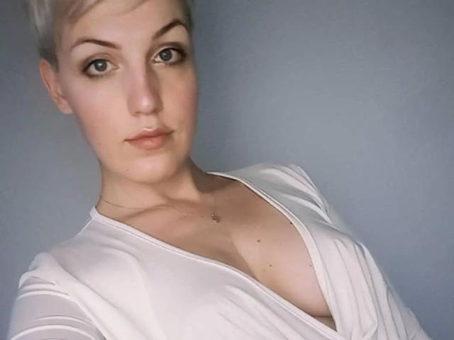 CharleneAngel-hot has short blonde hair, light skin, and small tits. She's wearing a white, low-cut top with a necklace, looking at the camera with a neutral expression.