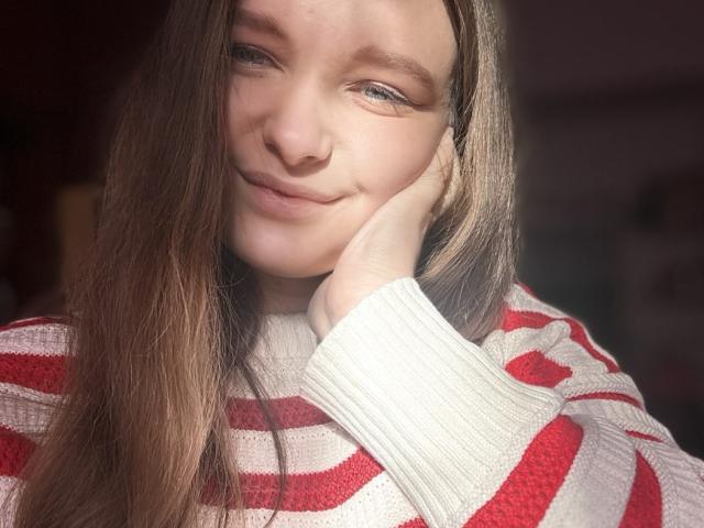 MagikPeachh has long, straight brown hair with a slight shine, and fair skin. She's wearing a red and white striped knit sweater. Her right hand rests on her cheek, and she's smiling softly with a slight tilt of her head. The light highlights her face, creating soft shadows. The texture of the sweater is ribbed, and the stripes are evenly spaced. Her blue eyes are slightly squinted, and her eyebrows are relaxed. The overall composition is warm and natural.
