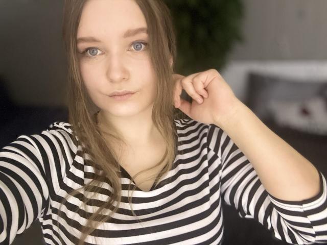 MagikPeachh has fair skin, blue eyes, and long, straight brown hair. She wears a black and white striped long-sleeve shirt. Her right hand is touching her hair. The image is a close-up, focusing on her face and upper body. The background is blurred, featuring a green plant and a dark, indistinct area. The lighting is even, highlighting her facial features and the texture of her shirt.
