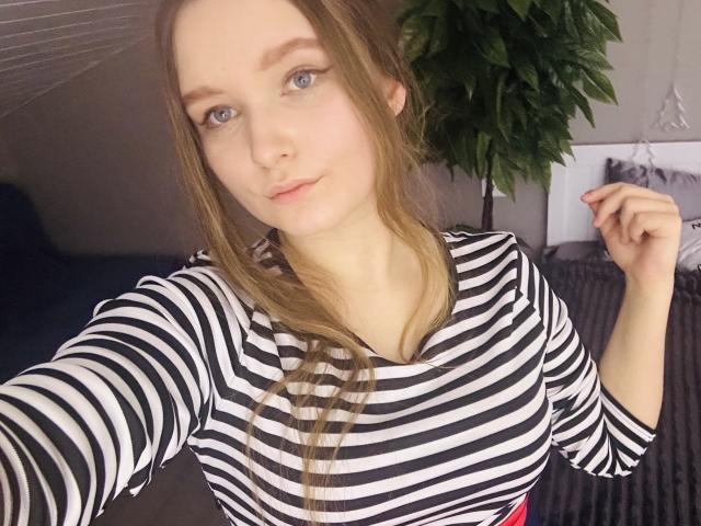MagikPeachh with long brown hair, blue eyes, and a striped black-and-white shirt, holding her hair, showing her small tits, in a selfie. She's cute and slightly smiling.