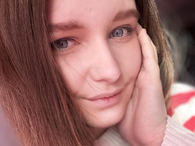 Close-up of MagikPeachh with light skin and straight brown hair. She has blue eyes and a subtle smile. Her right hand rests on her cheek. She's wearing a beige sweater. The image focuses on her face, highlighting her smooth skin and natural makeup. Her hair is slightly parted, covering part of her forehead. The background is blurred.