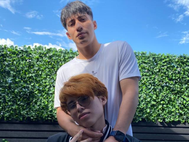 MaxAndTed with short brown hair and white tee, holding red-haired MaxAndTed with glasses and black wristband, standing against green hedge under blue sky.