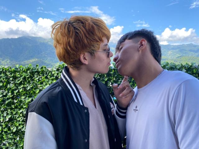 MaxAndTed with red hair and glasses, kissing nose of MaxAndTed with curly hair, wearing white shirt, black jacket, holding finger to chin, outside with green hedge.