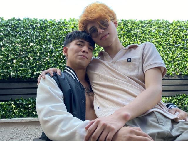 MaxAndTed with orange hair and glasses, wearing a beige polo, sits on the lap of MaxAndTed with black hair, wearing a black and white varsity jacket, outdoors.