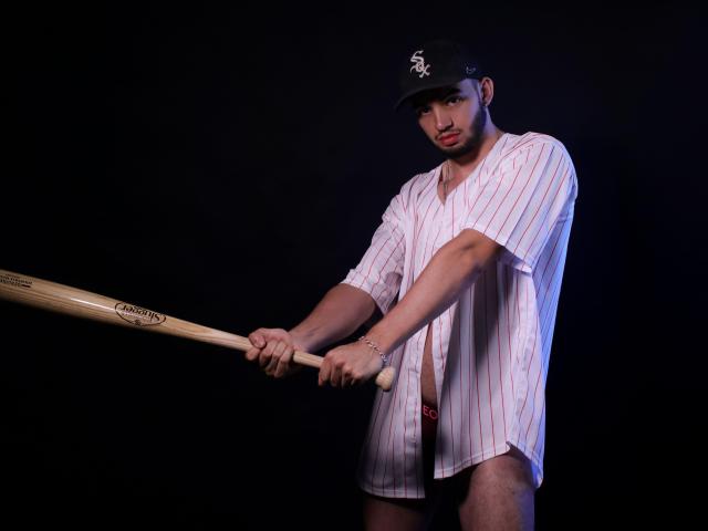 MichaelMagno stands against a dark background, holding a wooden baseball bat labeled "Easton." He wears a black cap with a white "N" logo, an oversized white baseball shirt with thin red stripes, and red underwear. His right hand grips the bat near the bottom, while his left hand holds it higher. MichaelMagno's serious expression contrasts with his relaxed posture. The lighting highlights his upper body, casting subtle shadows. The image focuses on his athletic build and the baseball gear.