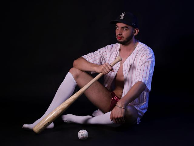 MichaelMagno sitting with a baseball bat, wearing a loose striped shirt, black cap, white socks, red underwear, and no pants, with a baseball on the floor.