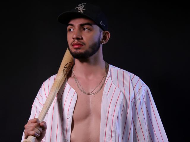 MichaelMagno with a baseball bat, wearing an open striped shirt, black cap, and chain necklace, has a beard, nose ring, and a serious look against a dark background.