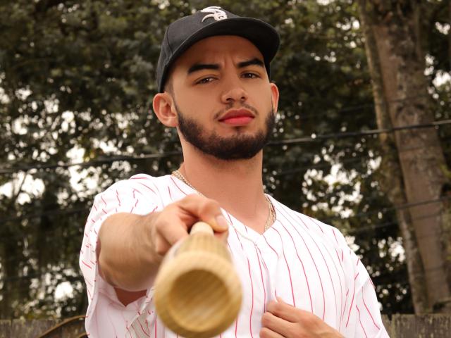 MichaelMagno with a beard, wearing a baseball cap and pinstripe shirt, pointing a wooden baseball bat towards the camera. He has a gold necklace and a slightly confident look.