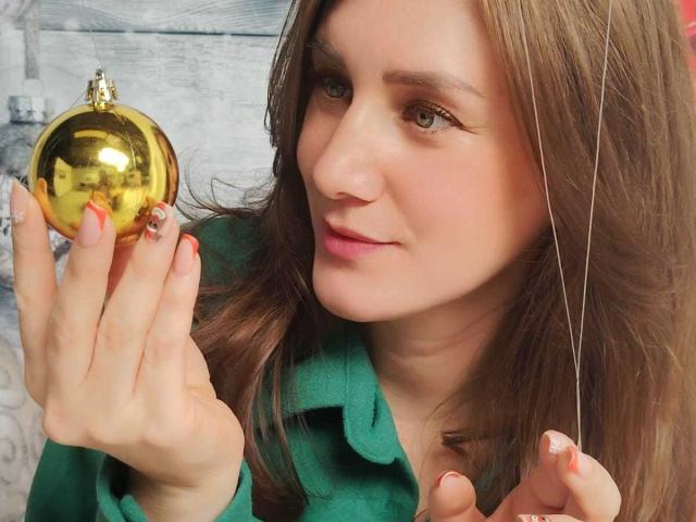 KatyaRogersX with brown hair, green eyes, and pink lips holds a shiny gold Christmas ornament, her pink-nailed hand near a thin string, wearing a green top.