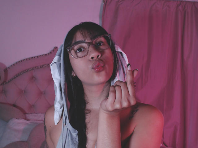 JessicaSoler with glasses, pouty lips, and gray headscarf, showing off her tits, holding her hand near her face, pink and purple lighting, tattoo on her right shoulder.