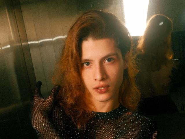 AllenClark with wavy red hair, wearing a sheer black polka dot top, has a serious expression, slightly parted lips, and a reflection of their head in a mirror behind.