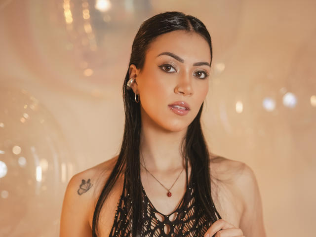 AbbieJonnes with straight black hair, wearing a black netted top, has a small tattoo on her right shoulder, and is looking directly at the camera with a neutral expression.