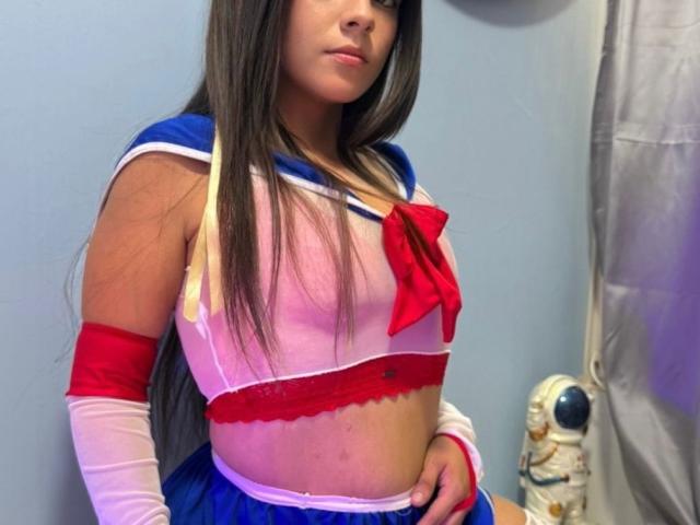 RilleyFoyx in a translucent pink and blue sailor outfit with red bow, showing her tits and belly, wearing red and white arm sleeves, long brown hair, and blue skirt.