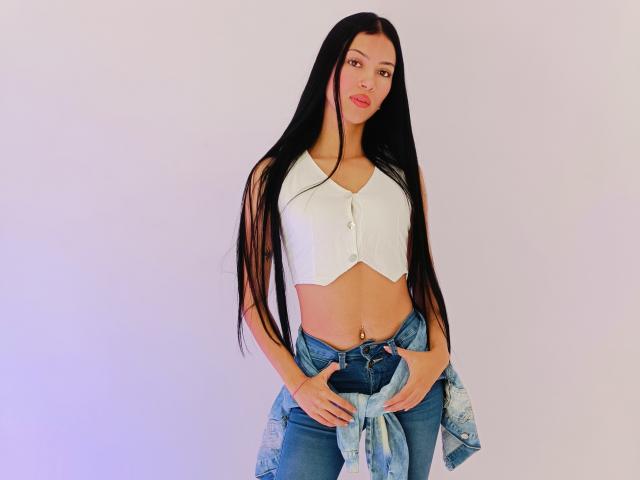 AbbieJonnes with long black hair, white cropped button-up, blue jeans, and a tied blue shirt, standing, showing a belly button piercing, looking at the camera with red lips.