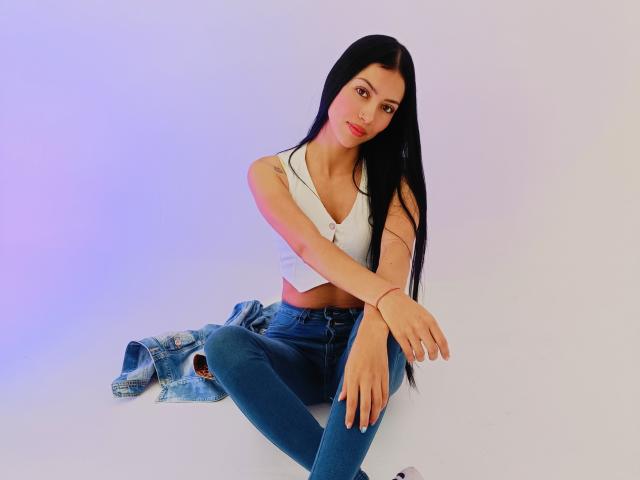 AbbieJonnes with long black hair, white crop top, and blue jeans, sitting with denim jacket on floor, arms crossed, slightly smiling, visible small tattoo on right shoulder.