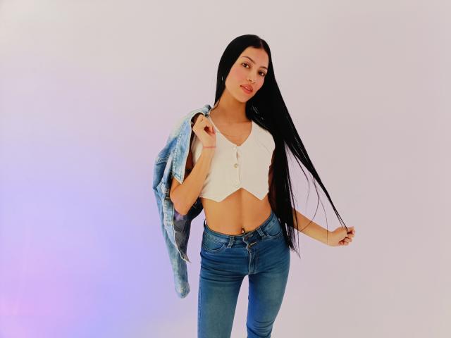 AbbieJonnes with long black hair, white crop top, blue jeans, holding a denim jacket, showing a belly button piercing, standing, smiling, and holding hair with her right hand.