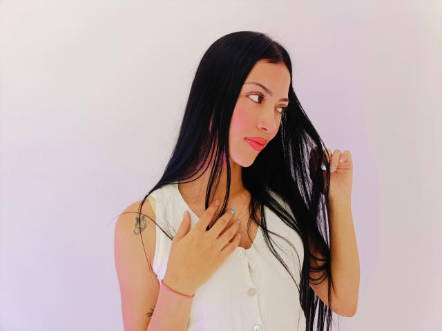 AbbieJonnes has long black hair, pink lipstick, and is wearing a white button-up shirt. She's touching her hair with one hand and her shirt with the other.