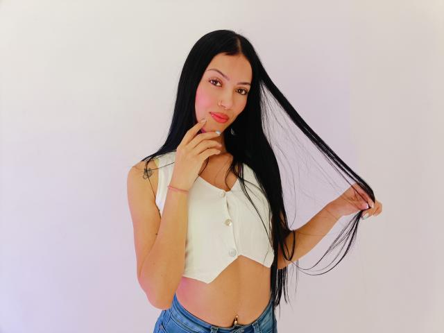 AbbieJonnes with long black hair, white crop top, showing small tits, and blue jeans, holding her hair, finger on lips, slight smile, light pink blush on cheeks.