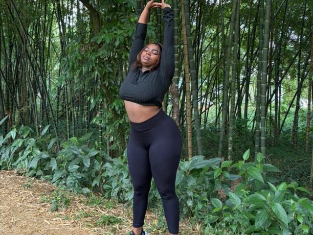 SamyLorenss stands in a forest, arms raised, eyes closed. She wears a black long-sleeve crop top and high-waisted black leggings, highlighting her curvy, full-figured physique. Her dark skin contrasts with the green foliage. The bamboo and leafy plants form a dense, vertical background. Her straight, dark hair is loose. The ground is covered in dry leaves and grass. The image has a natural, outdoor setting.