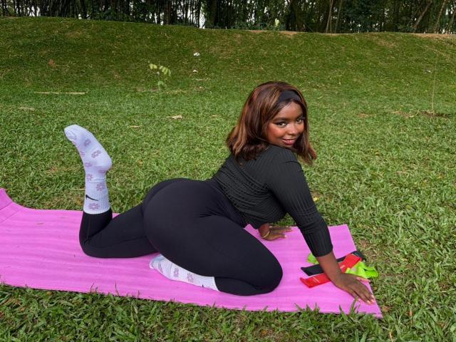 SamyLorenss on pink mat, black ribbed top, black pants, white socks, lying on grass, looking back, curvy ass, brown hair, smiling, outdoors, afternoon, green background.