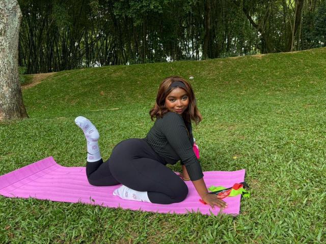 SamyLorenss is on a pink yoga mat in a grassy park, on all fours with her back to the camera. She has dark brown hair, a black headband, and is wearing a black long-sleeve top, black high-waist leggings, and white socks. Her right knee is bent with her foot up, and her left knee is on the mat. Her right arm is forward, and her left arm is bent with her hand on the mat. A yellow and red object is on the mat to her right.