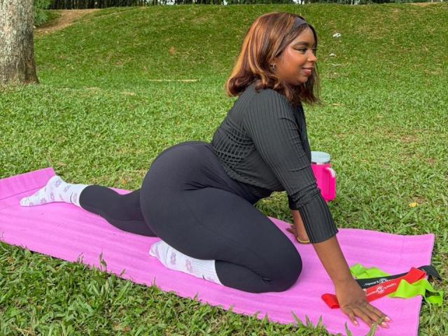 SamyLorenss is on a pink yoga mat in a grassy park, kneeling with one leg stretched out. She has dark brown, shoulder-length hair and is wearing a black rib-knit long-sleeve top and black leggings. Her socks are white with pink patterns. She's looking to the right with a slight smile. A red and green towel is on the mat to her right. The grass is green and there's a tree in the background. Natural light.