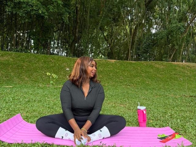SamyLorenss sits cross-legged on a pink mat in a grassy park. She wears a black rib-knit sweater, black pants, and white socks with colorful patterns. Her brown, wavy hair is loose. She smiles, looking to the right. A pink drink cup and a green and red book are on the mat. Trees with dense green foliage form the background. The composition is balanced with SamyLorenss centered and the mat's pink color contrasting with the green grass. The overall effect is casual and relaxed.