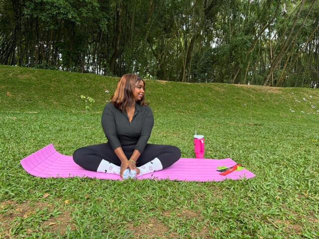 SamyLorenss sits cross-legged on a pink yoga mat in a grassy park. She's wearing a black hoodie, black pants, and white sneakers. Her brown hair is shoulder-length. She holds a blue water bottle in both hands. A pink water bottle and green and yellow pens are to her right. Green trees and grass form the background. The scene is outdoors with overcast lighting.