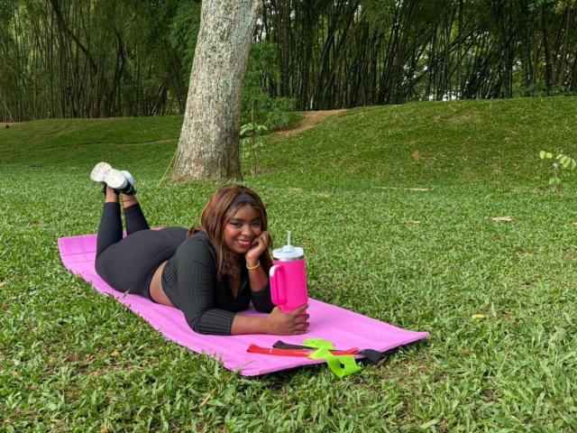 A woman, SamyLorenss, lies on a pink yoga mat in a grassy park. She has brown skin and brown hair, wearing a black crop top and black leggings. She holds a pink water bottle in her right hand, resting her head on her left hand. White sneakers are on her feet. A green and red object and a black object are near her. Trees and bamboo are in the background. Natural light. Casual, outdoor setting. Slightly smiling. SamyLorenss is in the foreground.
