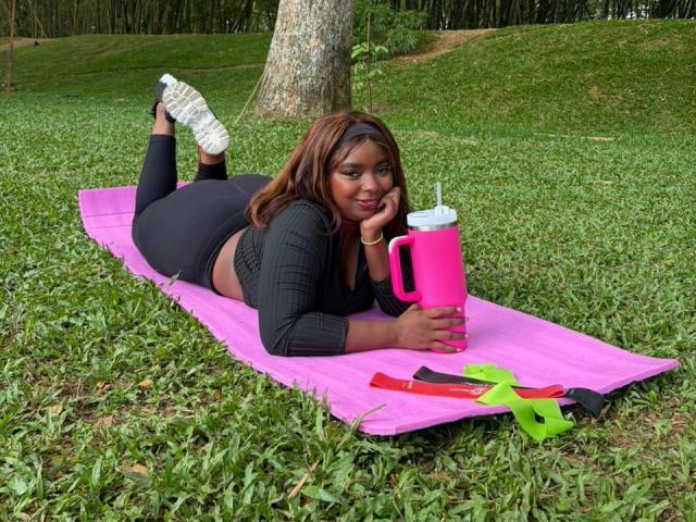 SamyLorenss lies on a pink yoga mat in a grassy park. She has dark brown skin and wears a black, long-sleeve crop top with black, tight-fitting pants. She holds a pink thermos with a white lid in her right hand. Her white sneakers are in the air. A green and red strap and a small green object are on the mat. SamyLorenss rests her chin on her left hand, smiling. The tree trunk is behind her.