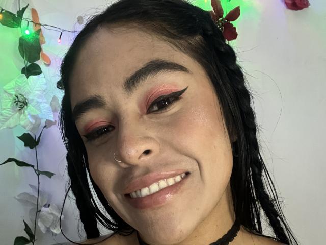 StellaBeckker has black hair in braids, pink and red eyeshadow, black eyeliner, a nose ring, and a smile showing white teeth. She's wearing a black choker.