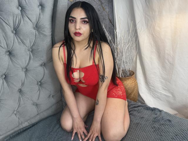 CamillaNoir with long black hair, red lace-up bodysuit, sitting on knees, gray tufted background, white curtain, and wicker basket with white branches, showing tattoos on arms.