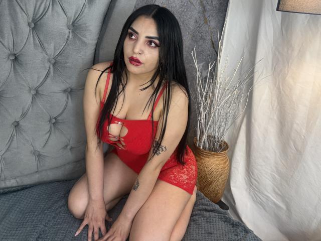 CamillaNoir with long black hair, red lace dress, and red lipstick, kneeling on a gray bed, showing her tits and tattoo, hands on bed, white branches in a basket.