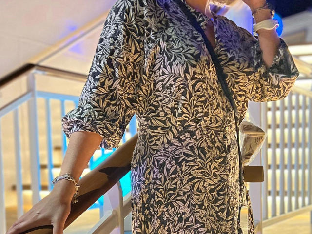 Zuckerschnecke-hot in a black and white floral pattern shirt, holding a drink in their right hand. The shirt has a loose, slightly rolled-up sleeve style. They wear a silver bracelet on their left wrist and a small crossbody bag. The background shows a brightly lit, modern staircase with a blue light. The image focuses on the upper body, with the face partially out of frame. The composition uses a slight diagonal angle.