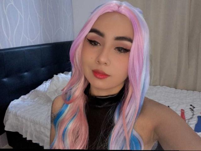 EmilyValk with pink, blue, and white hair, red lipstick, black top, and eyeliner, showing off her tits in a selfie. She's looking at the camera with a slight smile.