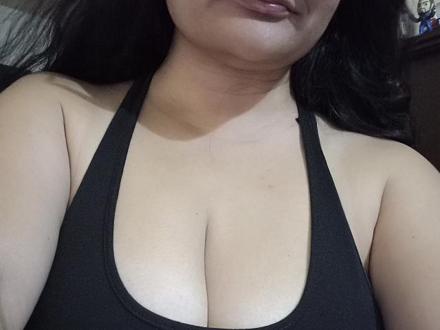 CamilaHot's large tits in a black halter top, showing a lot of cleavage with a slight tan and dark wavy hair, slightly parted glossy lips.