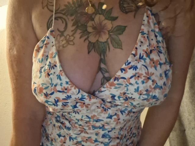 FrenchieLilieFleur's chest with large, colorful tattoos of flowers and leaves, wearing a low-cut, floral-patterned dress, showing her sizable, tattooed tits.