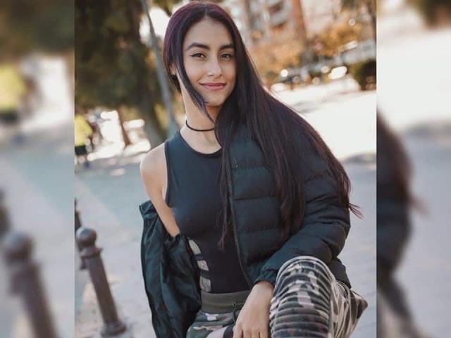 EmilySanz has long dark hair, a black puffy jacket, and black tank top with cut-out shoulders, smiling with her ass barely visible in camouflage pants.