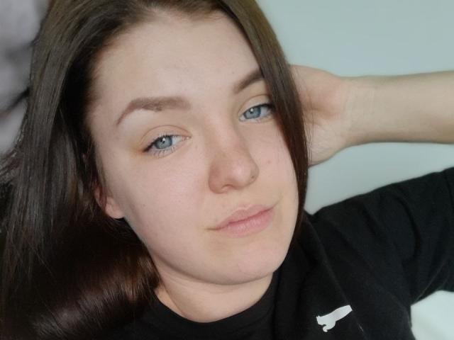MrsXSweet with blue eyes, brown hair, light skin, and a black shirt, resting her hand on the back of her head, looking slightly to the side with a subtle smile.