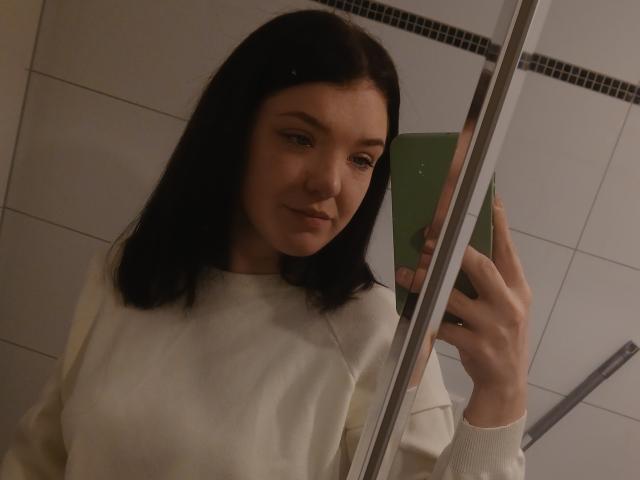 MrsXSweet with black hair, white sweater, holding green phone, taking mirror selfie, smiling slightly, in dimly lit bathroom. Boobs are small, face is attractive.