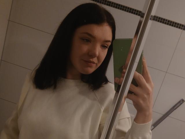 MrsXSweet with dark hair, white sweater, holding green phone, mirror selfie, subtle smile, soft lighting, visible nose, slightly parted lips, gentle expression, upper body shot.
