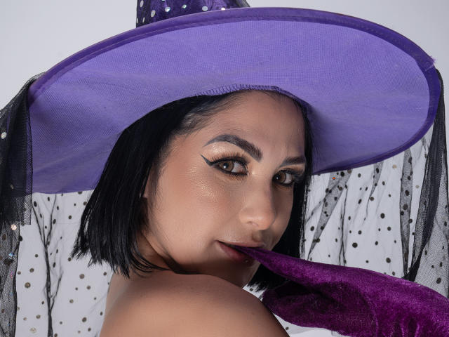 AntonellaFerreer with black bob, wearing purple witch hat, holding purple fabric in mouth, brown eyes with bold eyeliner, medium brown skin, looking seductively at camera.