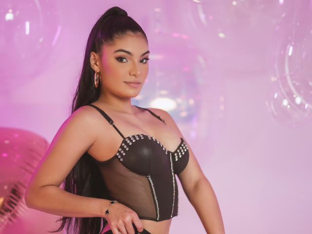 LunaAspen with long black ponytail, wearing a black mesh top with studded cups, looking at camera, pink lighting, earrings, confident smile, medium tits, cute, sexy, dark skin.