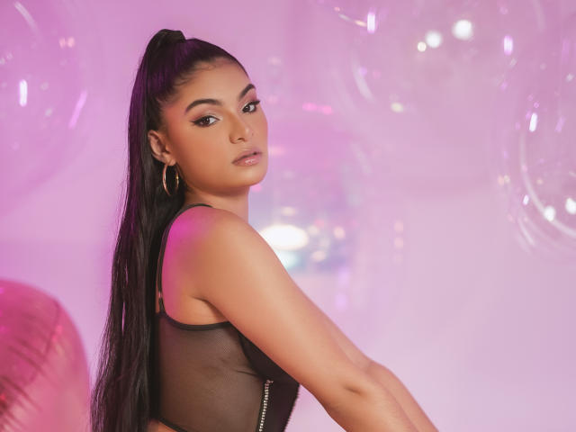 LunaAspen with long black hair in a ponytail, wearing a sheer black top, gold hoop earrings, looking over her shoulder with a serious expression, pink and purple lighting.