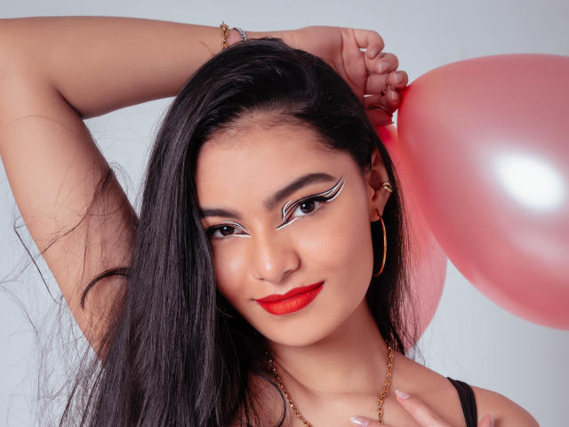 LunaAspen has long black hair, red lipstick, and white winged eyeliner. She holds a pink balloon, arm raised, wearing gold hoop earrings and a gold necklace.