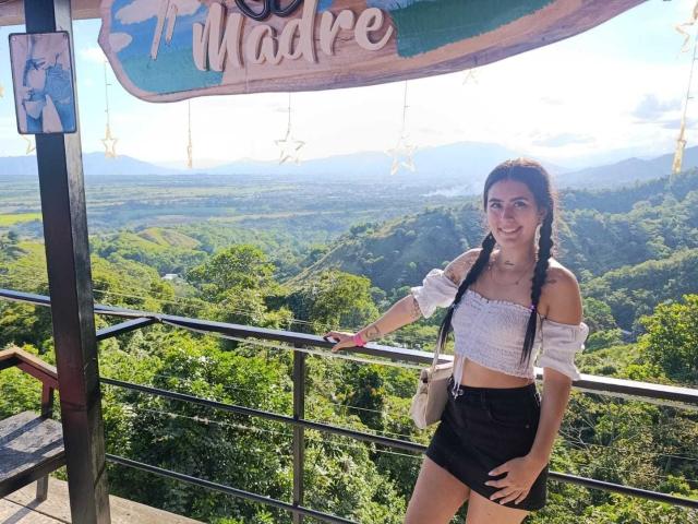 AlisonKweel stands at a viewpoint, right arm resting on a metal railing. She has long brown hair in two braids, wearing a white off-shoulder crop top and black shorts. She holds a smartphone in her left hand. Her white crossbody bag hangs over her right shoulder. The background shows a lush green valley with rolling hills and a clear sky. The sign above reads "Madre" with a star decoration. AlisonKweel smiles at the camera. Bright sunlight highlights her casual, summer outfit.