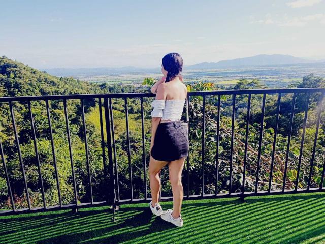 AlisonKweel stands on a green-striped balcony, facing away from the camera. She's wearing a white off-shoulder top and a black skirt with white sneakers. Her dark hair is down, and she's looking out over a lush, green landscape with rolling hills and distant mountains under a clear blue sky. The black rail and green balcony surface contrast with the natural scenery.