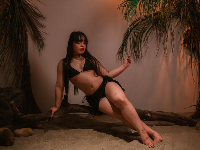 KateLeeh with long black hair, wearing a black bikini, sitting on a wooden log, legs crossed, palm leaves above, sand below, and warm orange lighting, showing her ass and tits.